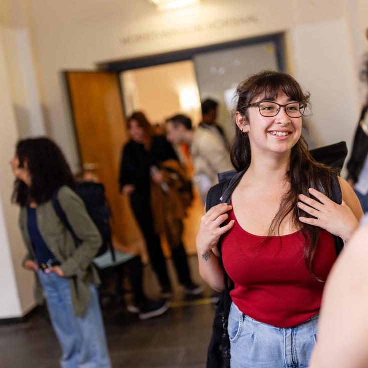 Mehrere Studierende stehen in einem Gang vor dem Hörsaal. Im Vordergrund unterhalten sich zwei Studentinnen.